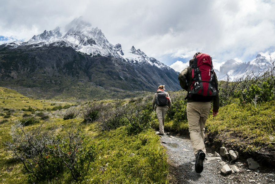Two hikers with backpacks walking a trail toward snow-capped mountains